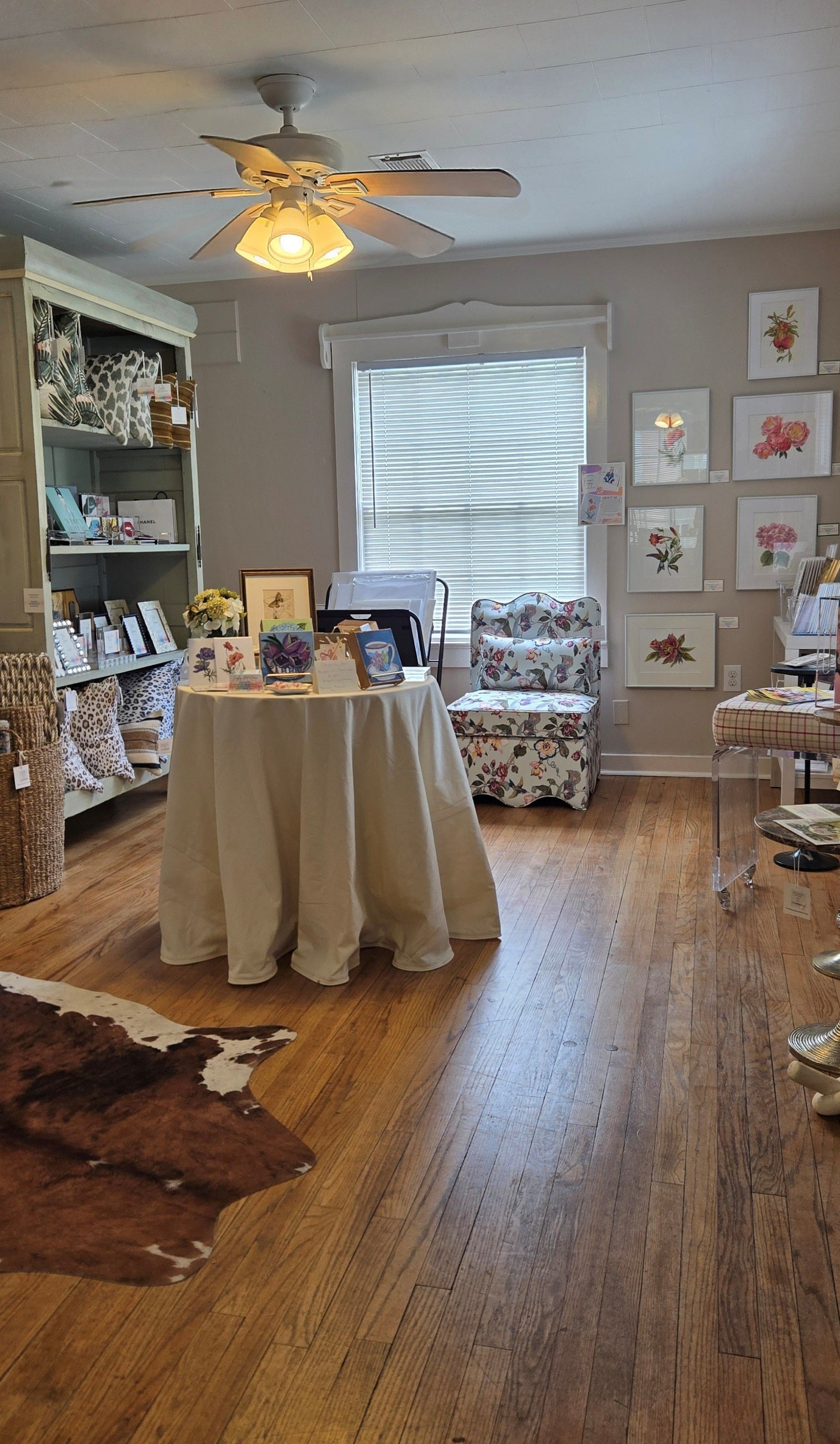 A sunlit gallery room at Langley Art & Home with framed botanical watercolors, a floral chair, and warm wood floors